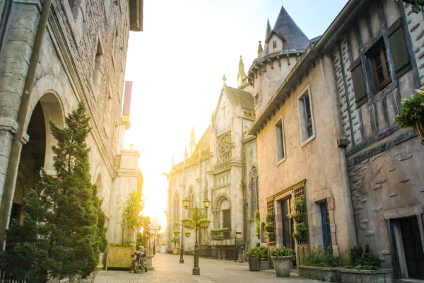 Charmante rue de style européen dans un village de château au coucher du soleil, avec des bâtiments en pierre, des tourelles et de la verdure.