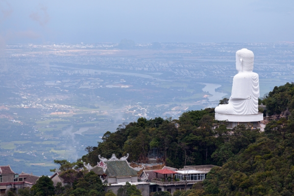 Grande statue de Bouddha blanche sur une colline surplombant une ville et une rivière, entourée d'une verdure luxuriante et de bâtiments traditionnels, idéale pour les voyages et le tourisme spirituel.