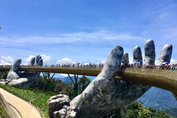 Pont d'or unique tenu par des mains de pierre géantes, entouré d'une végétation luxuriante et de montagnes sous un ciel bleu clair, une attraction touristique populaire au Vietnam.